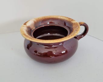 May include: A small, round, ceramic pot with a handle. The pot is a deep burgundy color with a lighter brown rim. The interior is dark brown. The pot is likely vintage or antique.