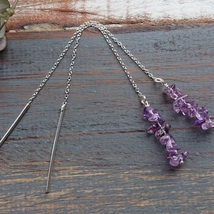 Puede incluir: Pendientes de hilo de plata con detalles de amatista. Los pendientes presentan largas y finas cadenas de plata, una barra en un extremo y un grupo de pequeñas piedras de amatista moradas en el otro.