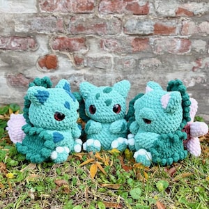 Può includere: Tre figure all'uncinetto fatte a mano nei toni del verde acqua e verde, che assomigliano a un popolare personaggio dei cartoni animati. Ogni figura ha gli occhi rossi ed è seduta sull'erba. Lo sfondo è un muro di mattoni.