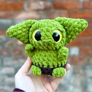 May include: A small, green, crocheted plush toy with large, glossy, yellow-rimmed black eyes and prominent pointed ears. The creature has a textured, yarn-based surface and a dark brown band around its base, resembling a simple garment.