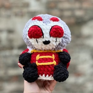 May include: A crocheted plush toy in the shape of a character with a grey head, red eyes, and a red and yellow outfit. The toy is sitting on a hand.