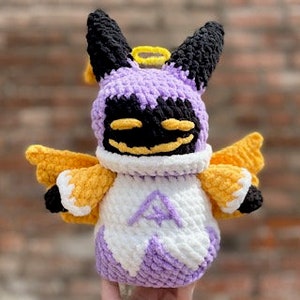 May include: A crocheted plush toy in the shape of a purple and black creature with yellow wings and a halo. The creature has a smiling face and is wearing a white robe.