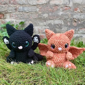 May include: Two crocheted plush toys shaped like dragons. One is black with white wings and the other is orange with pink wings. Both have large, round, blue eyes.