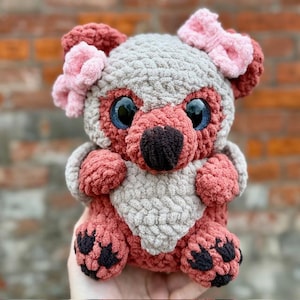 Può includere: Un peluche all'uncinetto fatto a mano a forma di orso. L'orso ha una testa grigia, orecchie e zampe rosso-marroni e un corpo rosso-marrone. Ha grandi occhi blu, un naso nero e un fiocco rosa. L'orso tiene in mano un petto grigio a forma di cuore.