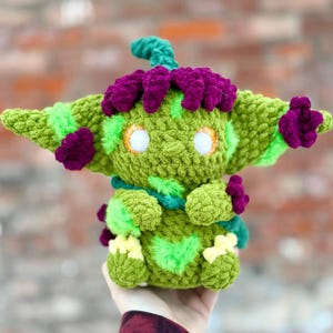Può includere: Una bambola all'uncinetto fatta a mano nei toni del verde, viola e giallo. La bambola presenta grandi orecchie, un copricapo e un disegno a forma di cuore sul petto. Gli occhi sono bianchi con accenti arancioni. La bambola è tenuta in una mano.