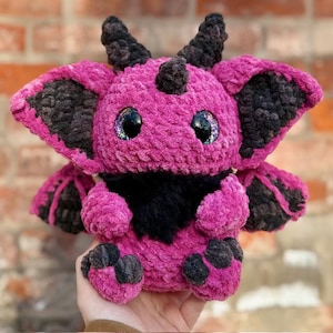 Puede incluir: Una criatura de peluche hecha a mano en tonos rosa y negro. El animal de peluche tiene ojos grandes y brillantes, cuernos pequeños y orejas grandes. La criatura tiene un pecho y almohadillas de patas de piel negra. El juguete de peluche está hecho de un material suave y texturizado.