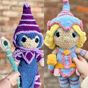 May include: Two crocheted amigurumi dolls, one in purple and blue with a wand, and the other in pink, blue and yellow with a yellow flower.