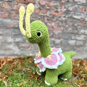 Può includere: Un dinosauro verde all'uncinetto fatto a mano con antenne gialle e un colletto decorativo di fiori rosa, bianchi e gialli. Il dinosauro è in piedi sull'erba con uno sfondo di muro di mattoni.