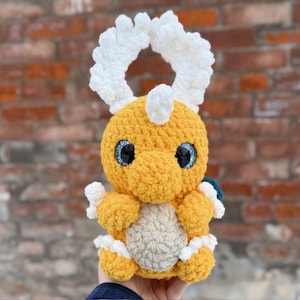 Puede incluir: Un juguete de peluche hecho a mano con forma de dragón de dibujos animados. El dragón es principalmente amarillo con detalles blancos, incluyendo un par de cuernos curvos y esponjosos. Tiene grandes ojos azules brillantes y una barriga gris. El juguete mide unos 15 cm de alto.