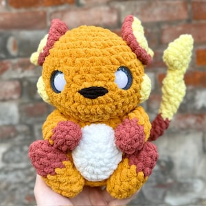 May include: A handmade, plush crochet creature in shades of orange, yellow, red, and white. The toy has large blue and white eyes, a black nose, and a tail. The creature is sitting with its arms and legs in front.