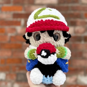 Può includere: Bambola all'uncinetto di un personaggio con un cappello bianco con bordo rosso e dettagli verdi. La bambola tiene in mano una palla rossa e bianca. Ha capelli neri, occhi marroni e una giacca blu.