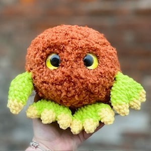 May include: A brown and green crocheted plush toy with large yellow eyes. The toy has a round body and four small green arms.