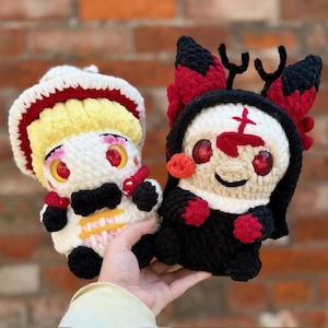 Op de afbeelding: Twee handgemaakte gehaakte poppen. De ene pop is wit, geel en rood, met een zwarte strik. De andere pop is zwart, wit en rood, met een kruis op het voorhoofd. Beide poppen hebben grote rode ogen en worden in een hand gehouden.