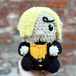 May include: A handmade crochet doll with a light yellow hairstyle, a yellow and black outfit, and large, expressive eyes. The doll's face is a light beige color, and it has a black mustache and eyebrows. The doll is held in a hand against a brick background.