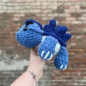May include: Blue and white crocheted plush toy resembling a cartoon dinosaur with a spiky back and large eyes.