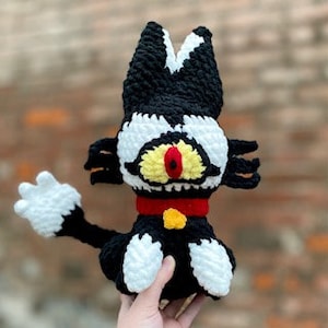 Può includere: Peluche di gatto in crochet nero e bianco con un collare rosso e un bottone giallo. Il gatto ha un grande occhio rosso.