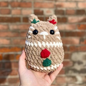 May include: A brown crocheted plush toy shaped like a gingerbread person with white, red, and green accents. The toy has a smiling face with black eyes and a red nose.