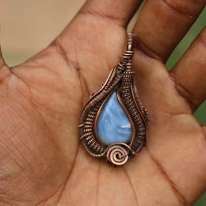 May include: A teardrop-shaped pendant featuring a smooth, light blue stone encased in intricate, dark copper wire wrapping. The metalwork includes detailed coils and woven patterns surrounding the central stone, with a small spiral at the base.