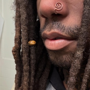 May include: A copper-coloured nose ring in a spiral design. The nose ring is worn on the left nostril. The person in the image has dark brown dreadlocks and a beard.