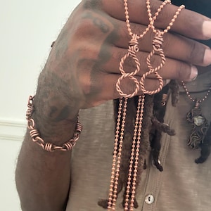 May include: A copper wire bracelet featuring a series of interconnected links wrapped with thin wire, and two matching copper pendants on ball chains, each designed with a twisted figure-eight shape and a wrapped wire bail at the top.