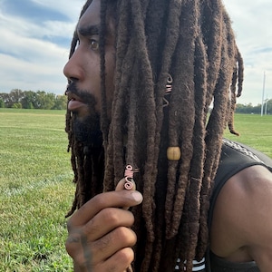 May include: A copper wire hair accessory featuring a spiral design, worn on a dark brown dreadlock. The accessory is made of coiled metal wire and is positioned on a single lock of hair, complementing a wooden bead on an adjacent dreadlock.