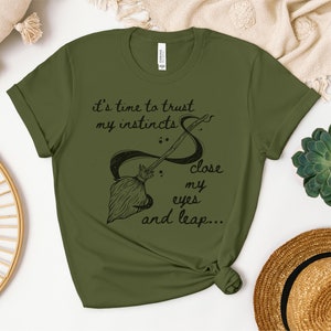 Könnte beinhalten: Olivengrünes T-Shirt mit einer schwarzen Grafik eines Besens und dem Text "it's time to trust my instincts & close my eyes and leap..."