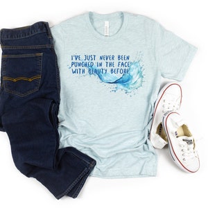 以下が含まれることがあります： 水色のTシャツに、水しぶきを上げる波の水彩画のデザインが施されています。Tシャツの文字は「I've just never been punched in the face with beauty before.」です。