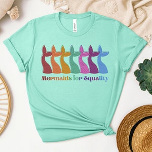 以下が含まれることがあります： 明るいティール色のTシャツ。カラフルな人魚の尾のイラストが並んでいます。尾は赤、オレンジ、黄色、緑、ピンク、紫、青の色合いです。「Mermaids for & equality」の文字が下に虹色でプリントされています。