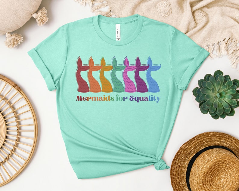 以下が含まれることがあります： 明るいティール色のTシャツ。カラフルな人魚の尾のイラストが並んでいます。尾は赤、オレンジ、黄色、緑、ピンク、紫、青の色合いです。「Mermaids for & equality」の文字が下に虹色でプリントされています。