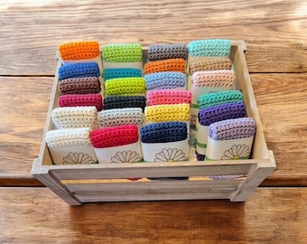 May include: A wooden crate filled with colorful, crocheted items. The items are in various colors, including orange, green, blue, and pink. Each item is displayed in a small white container with a daisy design. The crate sits on a wooden surface.