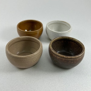 Puede incluir: Cuatro cuencos pequeños de cerámica en diferentes colores: marrón, beige, blanco y marrón con motas negras. Los cuencos son todos redondos y tienen un fondo ligeramente redondeado.