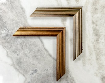 May include: Two picture frames, one gold and one silver, are shown against a white marble background. The frames are both ornate and have a raised edge.