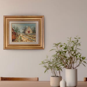 May include: A framed oil painting depicting a rural scene with a building and a path. The painting has a gold frame and is displayed on a neutral-colored wall. The scene includes a building with a red door and a path leading towards it.