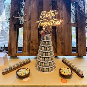 May include: A display featuring a macaron tower and decorative elements. The tower is surrounded by additional macarons and small bowls of candies. A wooden backdrop with the words "Better Together" in neon is visible.