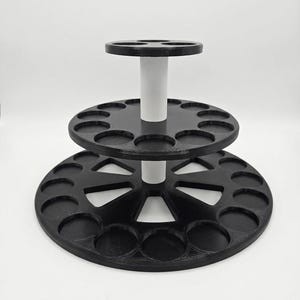 May include: A tiered circular display stand featuring a black base with multiple circular cutouts and a smaller black middle tier, both supported by a central white cylindrical post. The structure is designed to hold small round items.