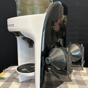 May include: A white and black baby formula maker with a clear container on top. The machine has a black base and a white top with the word "Babybrezza" printed on it. The machine has two black and gray dispensers on the side.