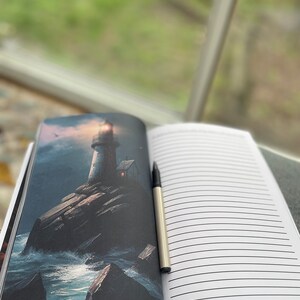 Op de afbeelding: Een open notitieboek met gelinieerde pagina's en een zilveren pen. De linkerpagina toont een donkere illustratie van een vuurtoren aan een rotsachtige kust met golven die tegen de rotsen beuken.