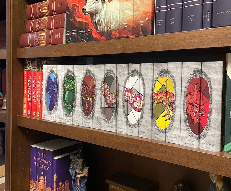 May include: A bookshelf displays a collection of fantasy books. The spines of the books feature colorful, stained-glass-style dragon designs. Other books on the shelf have burgundy and navy blue covers. The books are arranged on a wooden bookshelf.