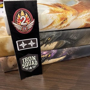 May include: A black leather bookmark with three patches. The top patch is red, white, and yellow with the number "2" and the Roman numeral "IV". The middle patch is white with two stars. The bottom patch is black with white text that reads "IRON SQUAD" and a white star.