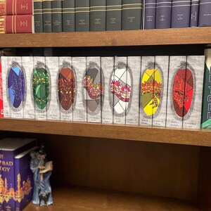 May include: A wooden bookshelf filled with books. The top shelf holds hardcover books in various colors. The middle shelf displays a series of books with colorful, stained-glass-like designs. A purple box set and a figurine are on the bottom shelf.