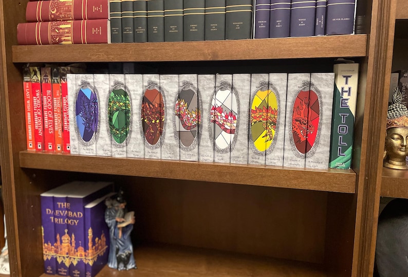 May include: A wooden bookshelf filled with books. The top shelf holds hardcover books in various colors. The middle shelf displays a series of books with colorful, stained-glass-like designs. A purple box set and a figurine are on the bottom shelf.