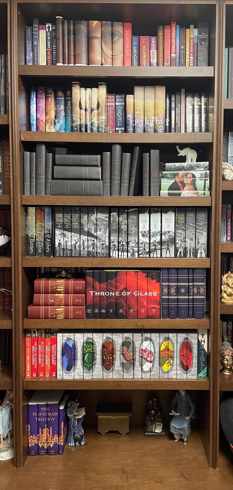 May include: A wooden bookshelf filled with various books. The books are of different sizes and colors, with some featuring the text "THRONE OF GLASS" and "THE DAEVABAD TRILOGY". Decorative items are also present.