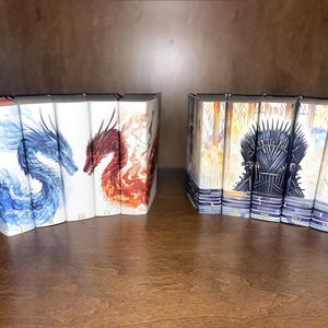 May include: A set of nine hardcover books with illustrated covers. The covers feature a blue and white dragon, a red and orange dragon, and a black iron throne. The books are arranged in two rows on a wooden shelf.