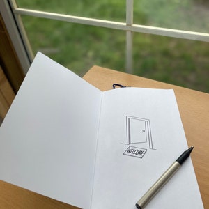 Op de afbeelding: Een open notitieboek met een zwarte pen erop. Het notitieboek heeft een blauw lint als bladwijzer en een eenvoudige tekening van een open deur met een welkomstmat.