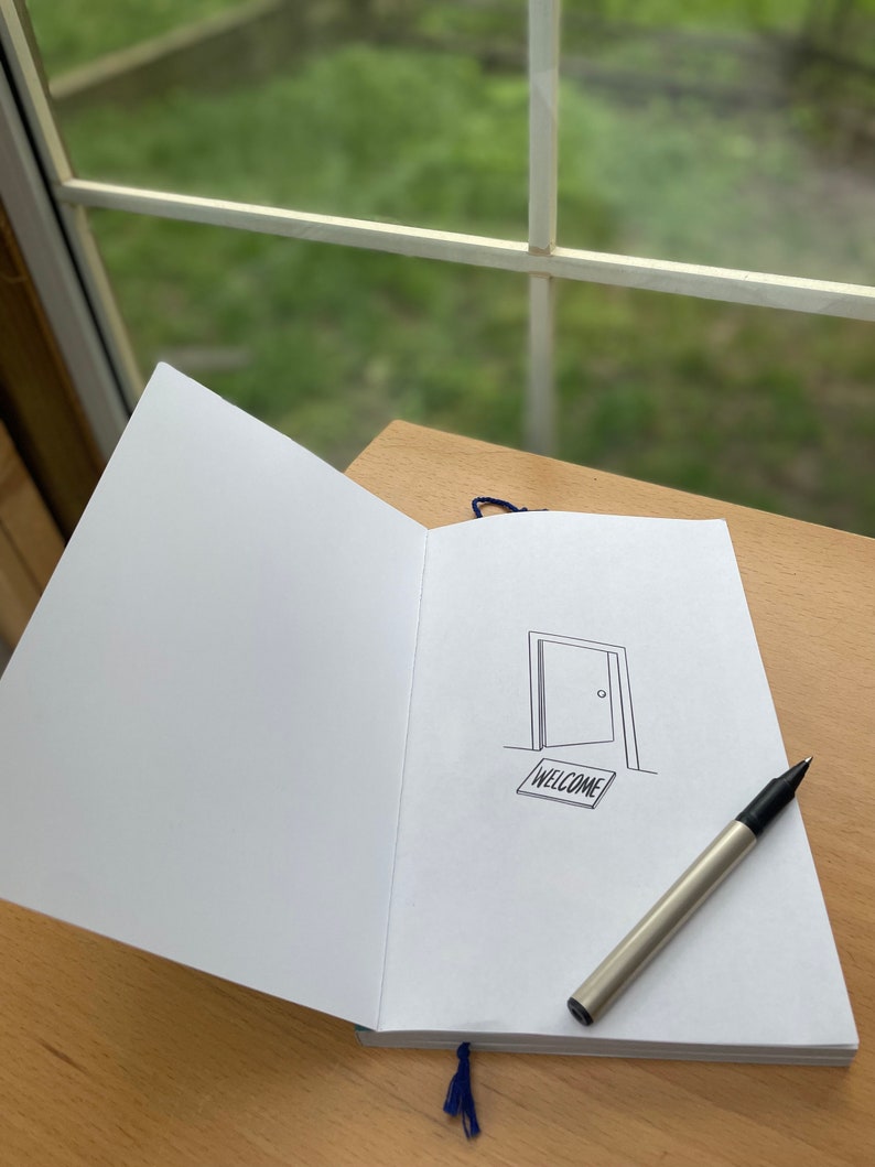 Op de afbeelding: Een open notitieboek met een zwarte pen erop. Het notitieboek heeft een blauw lint als bladwijzer en een eenvoudige tekening van een open deur met een welkomstmat.