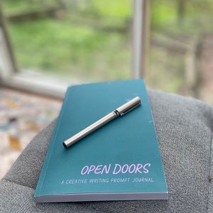 Op de afbeelding: Een turquoiseblauw notitieboekje met de tekst "OPEN DOORS A CREATIVE WRITING PROMPT JOURNAL" op de omslag. Een zilveren pen ligt op het notitieboekje.