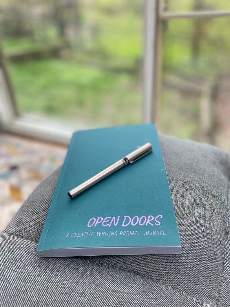Op de afbeelding: Een turquoiseblauw notitieboekje met de tekst "OPEN DOORS A CREATIVE WRITING PROMPT JOURNAL" op de omslag. Een zilveren pen ligt op het notitieboekje.