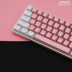 May include: A white and pink mechanical keyboard with a pink space bar and keycaps. The keys are labeled with the standard alphabet and number keys. The keyboard is on a pink surface.