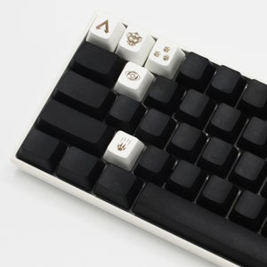 May include: A black mechanical keyboard with white keycaps featuring various symbols, including a triangle, an eye, and paw prints. The keyboard has a black space bar and a white frame.