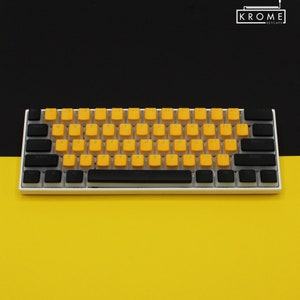 May include: A white mechanical keyboard with black keycaps and yellow keycaps. The keyboard has a black space bar and a black frame. The keyboard is on a yellow surface.